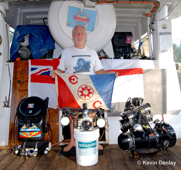 Kevin with Explorers Club Flag #46 and the ‘tools of his trade’ aboard MV Empress; left, his MK15.5 Closed Circuit Rebreather; centre, his housed Nikon digital camera atop a tub of Co2 absorbent for his CCR; right his Diver Propulsion Vehicle, or scooter, with a housed digital video camera mounted atop.Note Empress’s on-board recompression chamber behind Kevin’s head