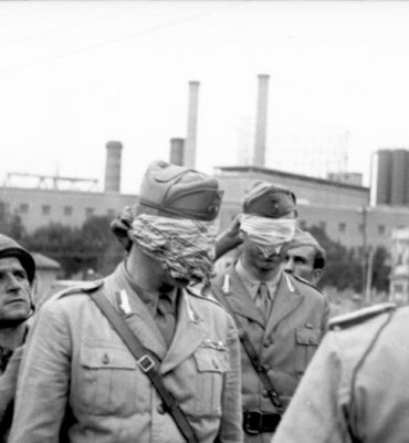 This image is referred to the surrender of the city of Rome. The two italian officers are part of the italian delegation. They have been blindfolded by the fallschirmjager that will take them to the german headquarters. At that time they were not prisoners yet.