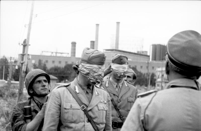 This image is referred to the surrender of the city of Rome. The two italian officers are part of the italian delegation. They have been blindfolded by the fallschirmjager that will take them to the german headquarters. At that time they were not prisoners yet.